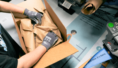 Hands are shown placing packaging material inside a cardboard box. 