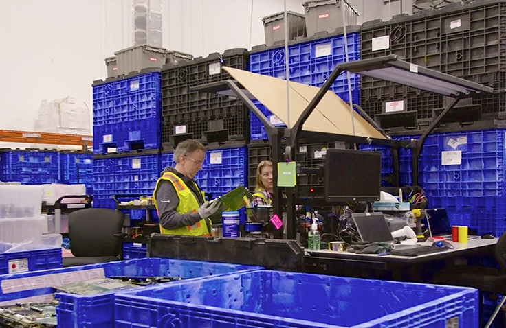 Two people surrounded by black and blue bins work in front of a large piece of machinery. 