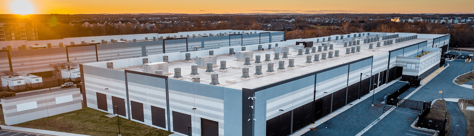 Aerial view of a large data center at sunset, featuring a modern design with a gray and black exterior. The building has multiple ventilation units on the roof and is surrounded by a paved parking lot. In the background, the sun sets over a distant landscape, casting warm colors in the sky.