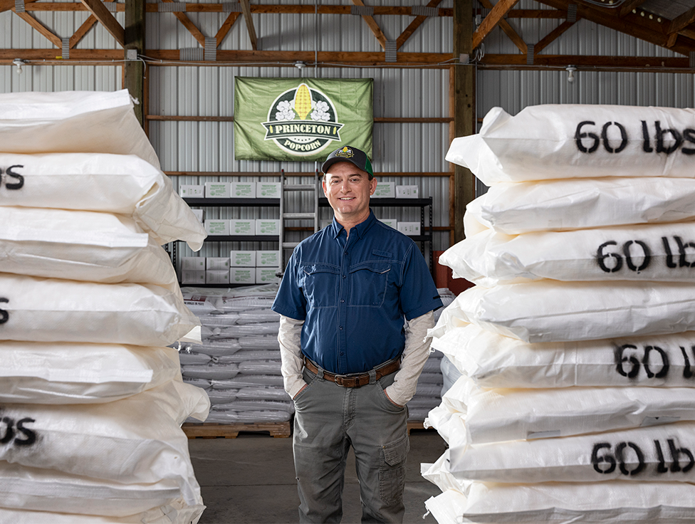 A person stands smiling in between 60 lb bags of product, with a sign in the back that says: "Princeton Popcorn".