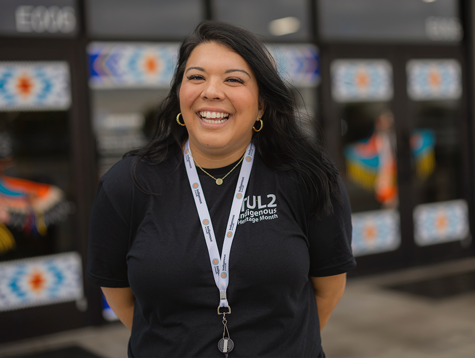 A person wearing a lanyard and a t-shirt that says "indigenous heritage month" stands smiling.