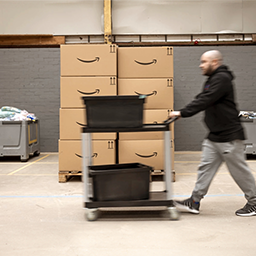 An employee is shown pushing a cart past two stacks of cardboard Amazon boxes.